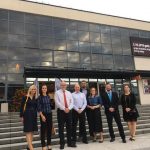 Grupa osób przed budynkiem z przeszkloną fasadą i banerem wydarzenia.