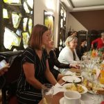 Grupa ludzi przy stole pełnym potraw i napojów na przyjęciu w elegancko urządzonym wnętrzu.