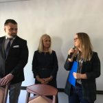 Trzy osoby przy biurku w formalnych strojach, jedna mówi do mikrofonu.